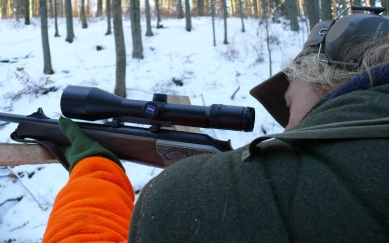 Ein Schütze in grün-orangefarbener Kleidung, von dem nur Kopf und Schulter zu erkennen sind, blickt in einem winterlichen Wald durchs Zielfernrohr seiner Waffe