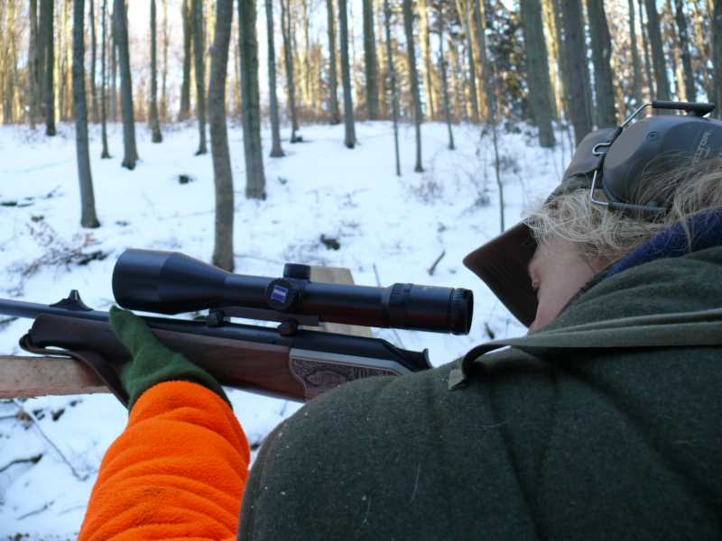 Ein Schütze in grün-orangefarbener Kleidung, von dem nur Kopf und Schulter zu erkennen sind, blickt in einem winterlichen Wald durchs Zielfernrohr seiner Waffe