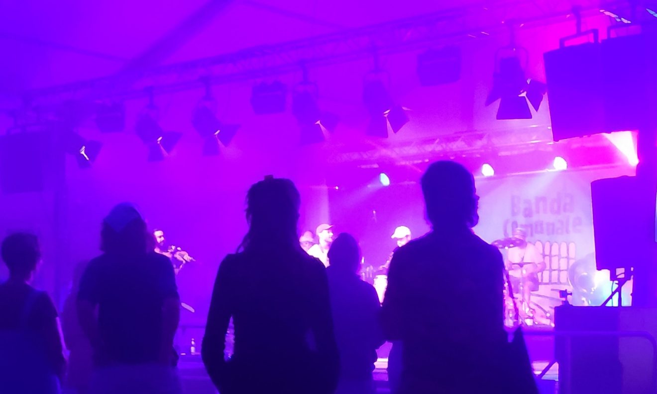 Mehrere Menschen stehen vor einer pinkfarben beleuchteten Bühne, auf der eine Band spielt