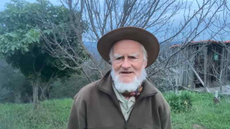 Ein älterer Mann mit weißem Bart und braunem Hut vor Naturlandschaft