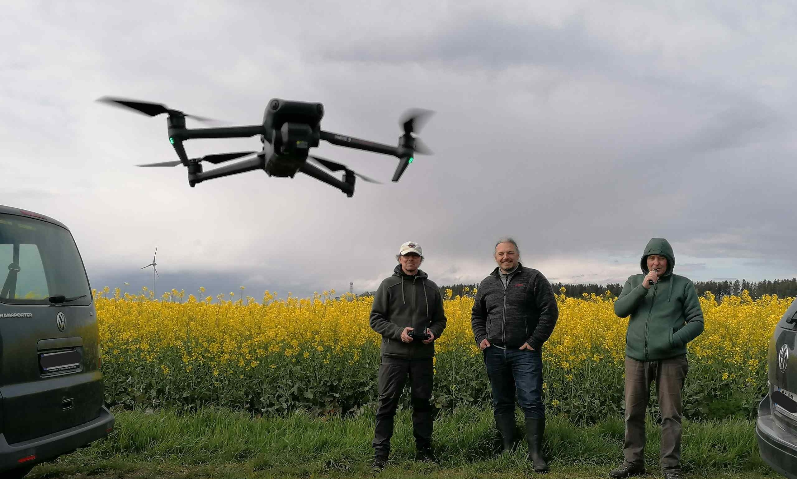 Drei Personen stehen am Feldrand zwischen Autos, vor ihnen schwebt eine Drohne auf den Betrachter zu