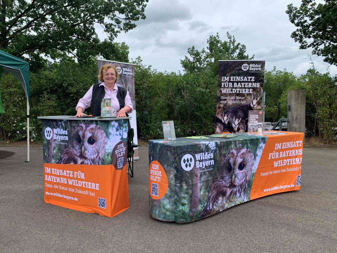 Auf einem Platz vor Bäumen ist ein Messestand von Wildes Bayern aufgestellt, eine Frau steht dahinter