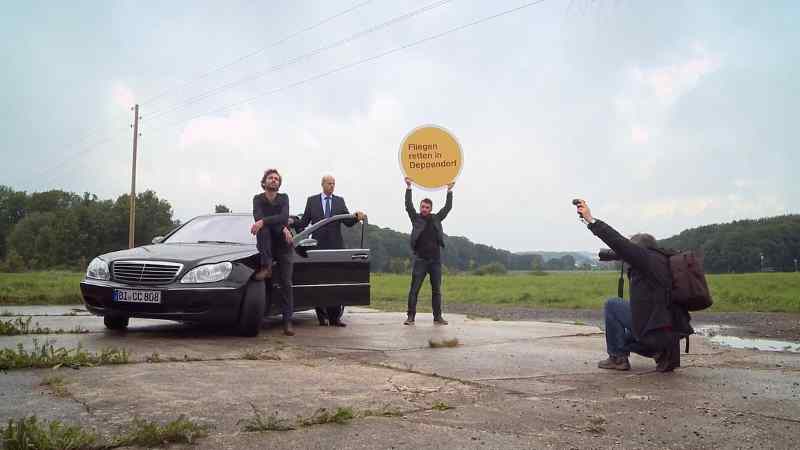 An einem Auto lehnen zwei Männer, ein dritter hält ein Schild "Fliegen retten in Deppendorf" hoch, ein vierter fotografiert