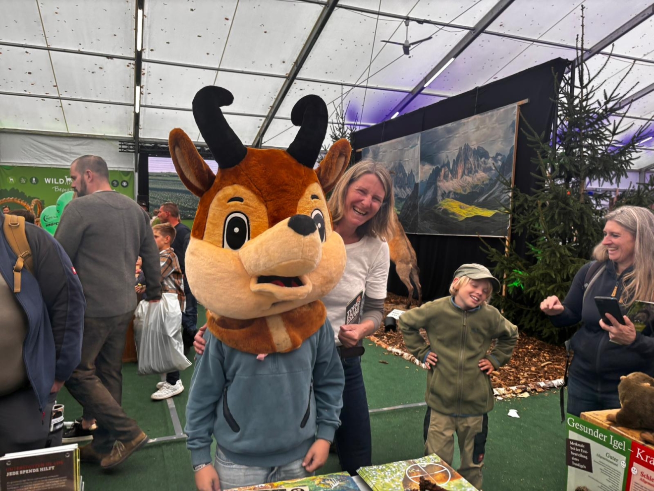 Das Foto zeigt ein Gams-Maskottchen auf einer Messe, hinter dem eine blonde Frau hervor schaut, daneben ein Kind