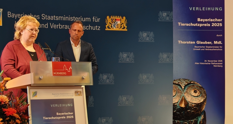 Vor blauem HIntergrund steht eine blonde Frau mit Brille und roter Bluse an einem Rednerpult, im Hintergrund steht ein Mann