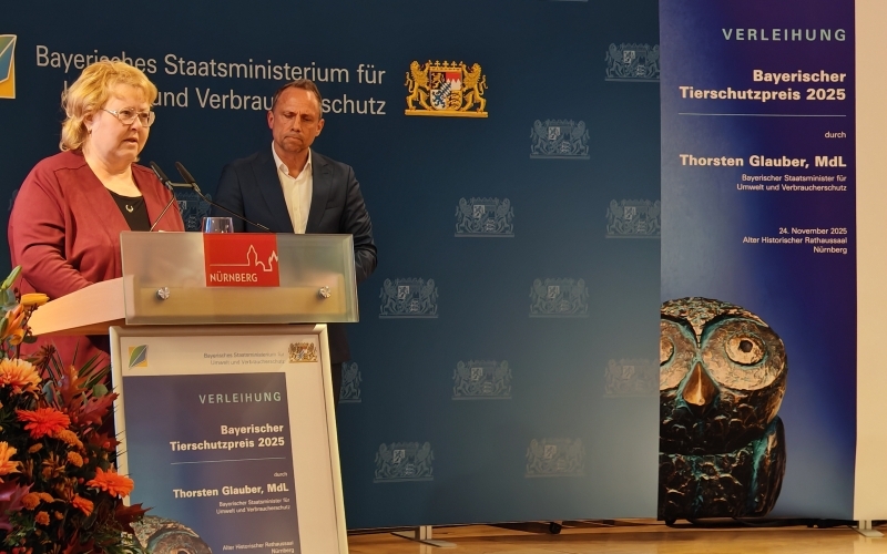 Vor blauem HIntergrund steht eine blonde Frau mit Brille und roter Bluse an einem Rednerpult, im Hintergrund steht ein Mann