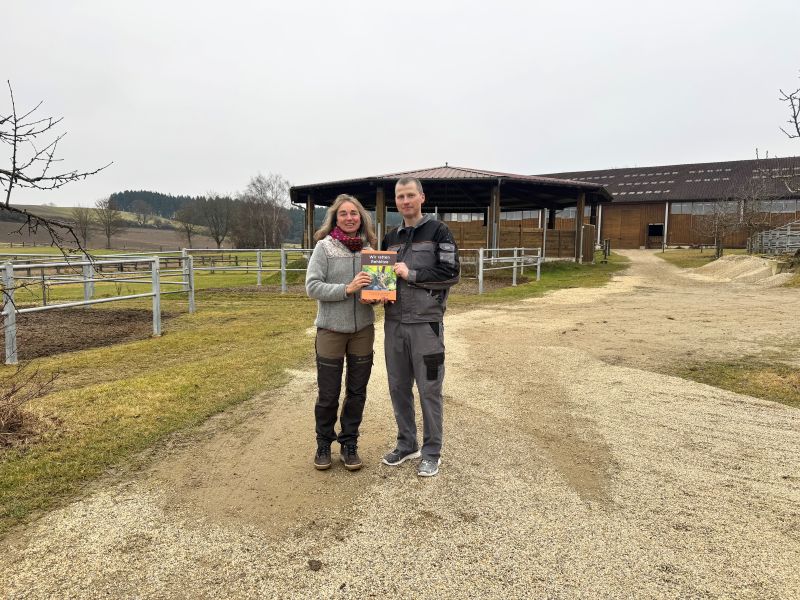 Ein Mann und eine Frau stehen vor einer Pferde-Reitanlage, sie halten gemeinsam ein Buch in die Kamera.