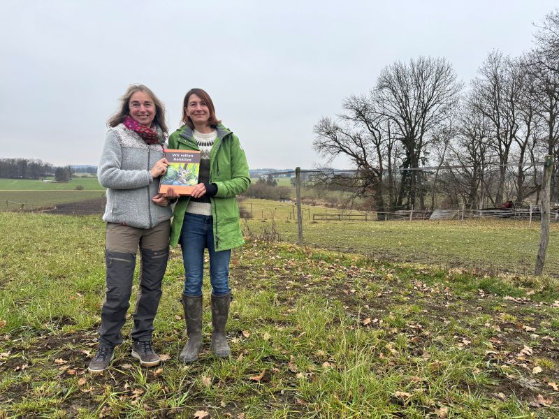 Zwei Frauen stehen vor einer offenen Graslandschaft, sie halten gemeinsam ein Buch in die Kamera.