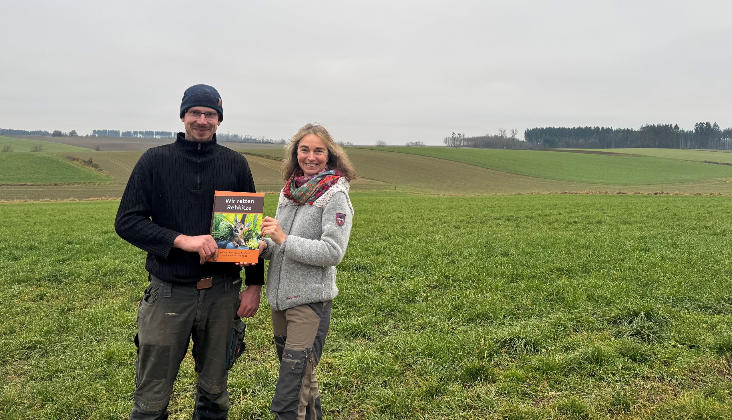 Ein Mann und eine Frau stehen vor einer offenen Graslandschaft, sie halten gemeinsam ein Buch in die Kamera.