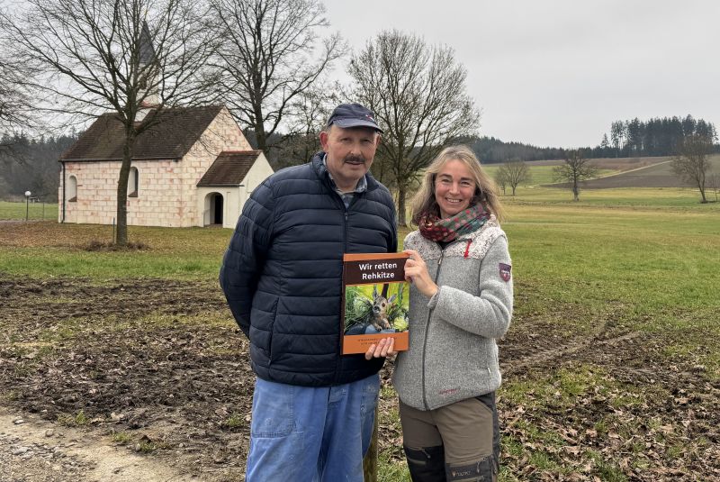 Ein Mann und eine Frau stehen in einer Landschaft, sie hält zwischen ihnen ein Buch in die Kamera.