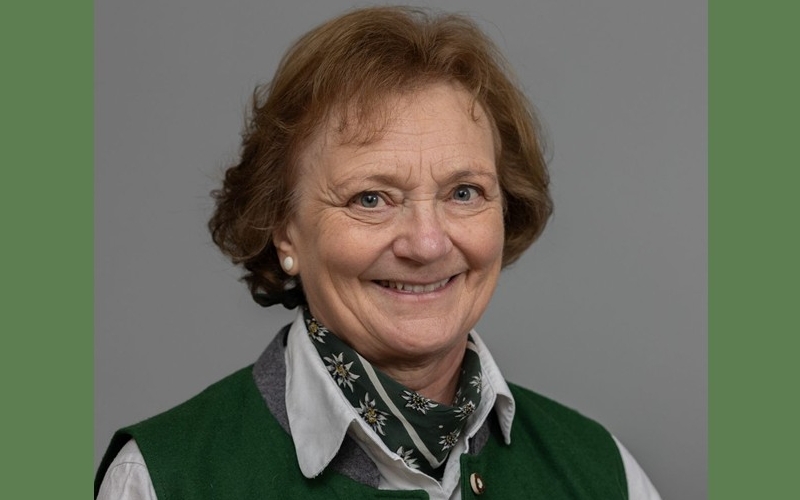 Vor grünem Hintergrund das Porträtfoto einer älteren Frau mit kurzen braunen Locken, Bluse und Trachtenhalstuch