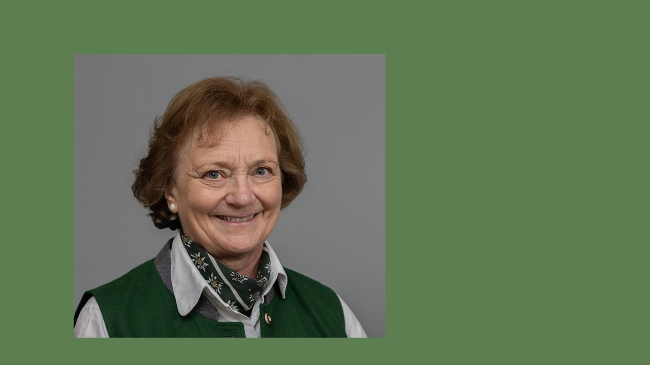 Vor grünem Hintergrund das Porträtfoto einer älteren Frau mit kurzen braunen Locken, Bluse und Trachtenhalstuch