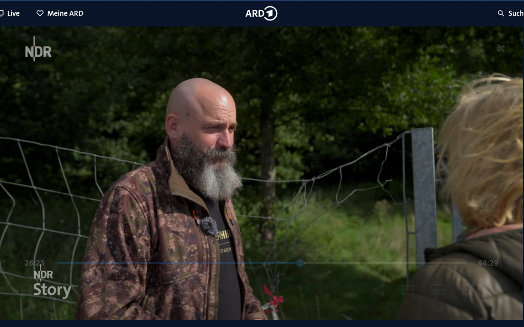 Screenshot einer Filmszene, in der ein Mann in Tarnkleidung mit Bart vor grünem Hintergrund mit einer Frau spricht.