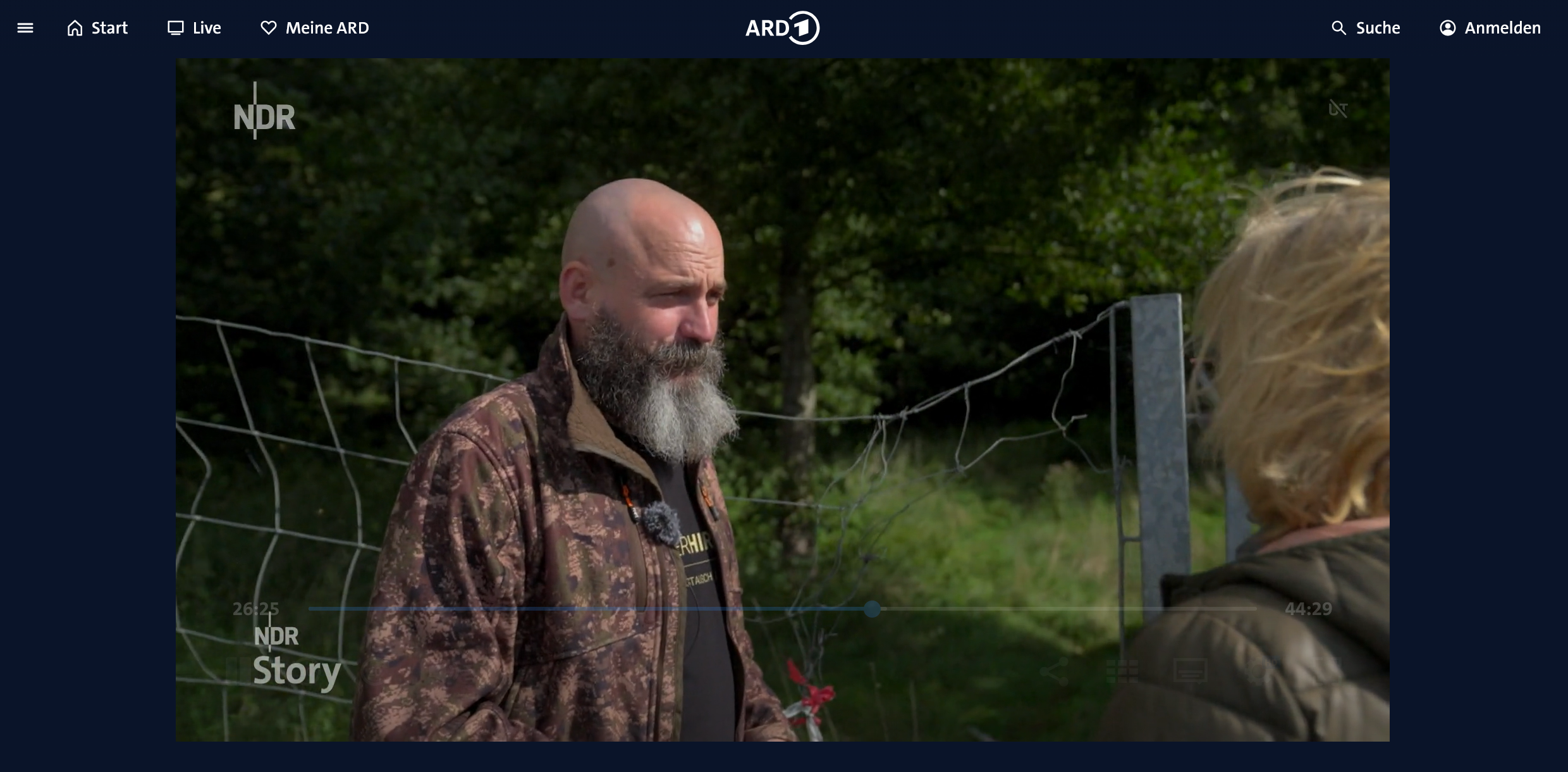 Screenshot einer Filmszene, in der ein Mann in Tarnkleidung mit Bart vor grünem Hintergrund mit einer Frau spricht.