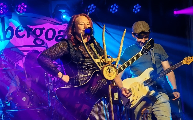 Ein Mann mit Gitarre und eine Frau als Musiker auf einer Bühne, türkis-blaues und violettes Licht