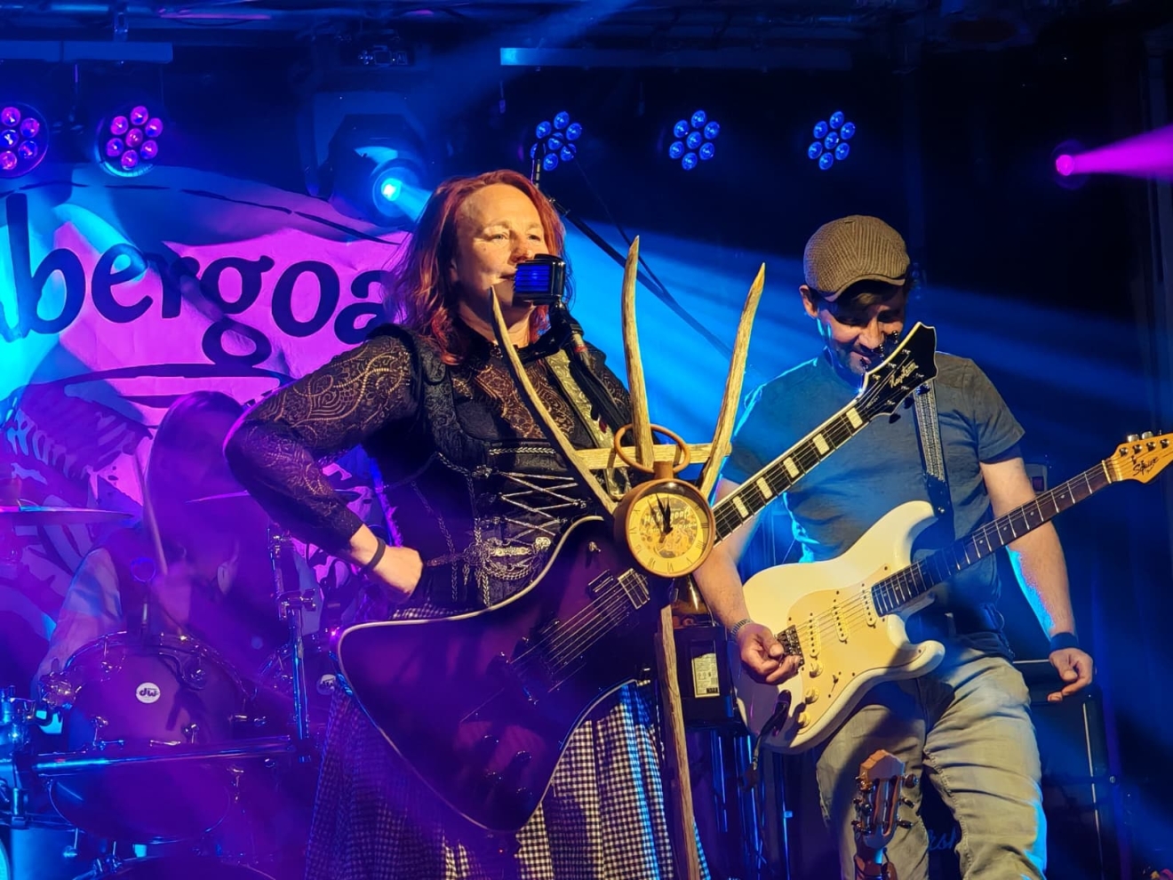 Ein Mann mit Gitarre und eine Frau als Musiker auf einer Bühne, türkis-blaues und violettes Licht