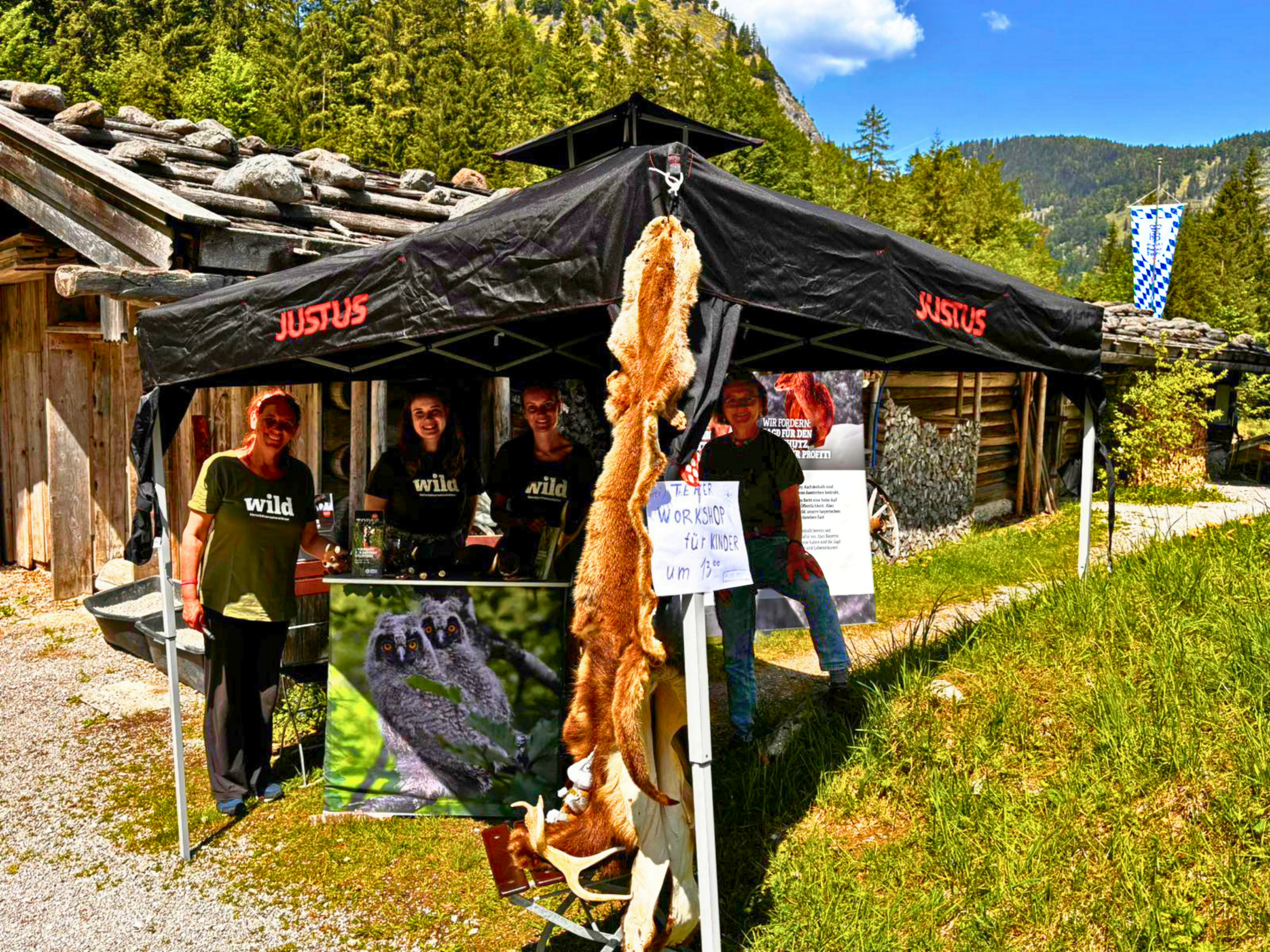 Vor einer sommerlichen Gebirgskulisse steht neben einer Holzhütte ein schwarzer Pavillon, an dem ein Tierfell hängt, und unter dem vier junge Menschen mit "Wild"-T-Shirts stehen