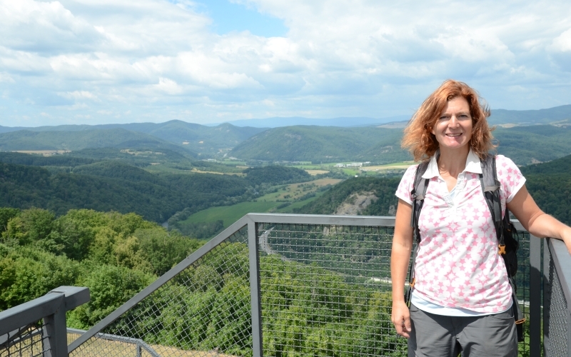 Eine Frau mit Rucksack lehnt an einem Geländer, dahinter Landschaftsausblick auf bewaldete Hügel und einen Fluss