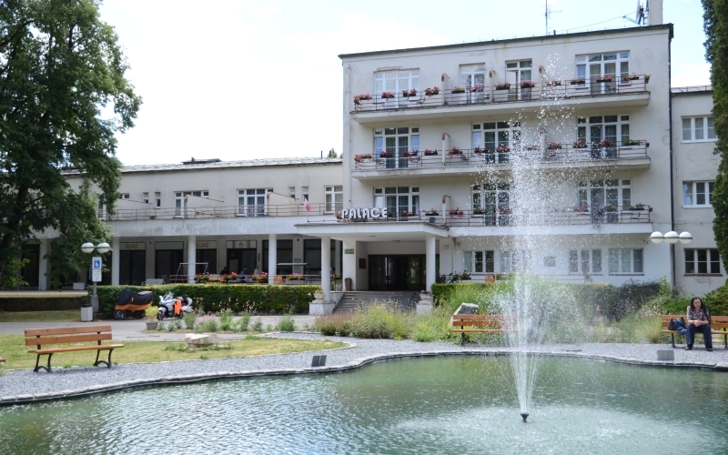 Im Vordergrund ein Teich mit Springbrunnen, dahinter Blumenrabatten und ein sozialistisch anmutendes, mehrstöckiges helles Gebäude mit der Aufschrift "Palace"