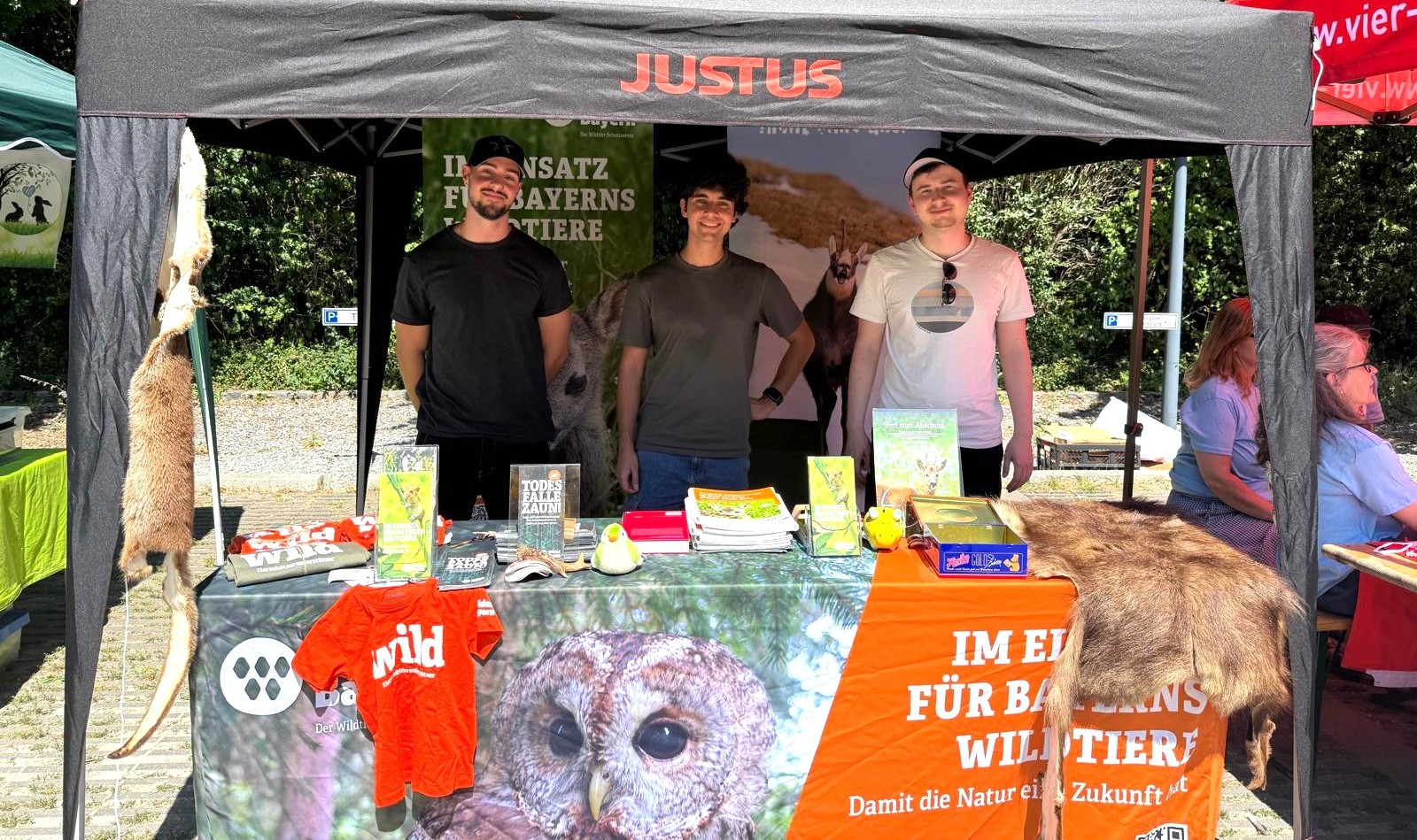 Ein Messestand im Freien unter einem Pavillon mit Produkten und Aufstellern von Wildes Bayern, drin stehen drei junge Männer
