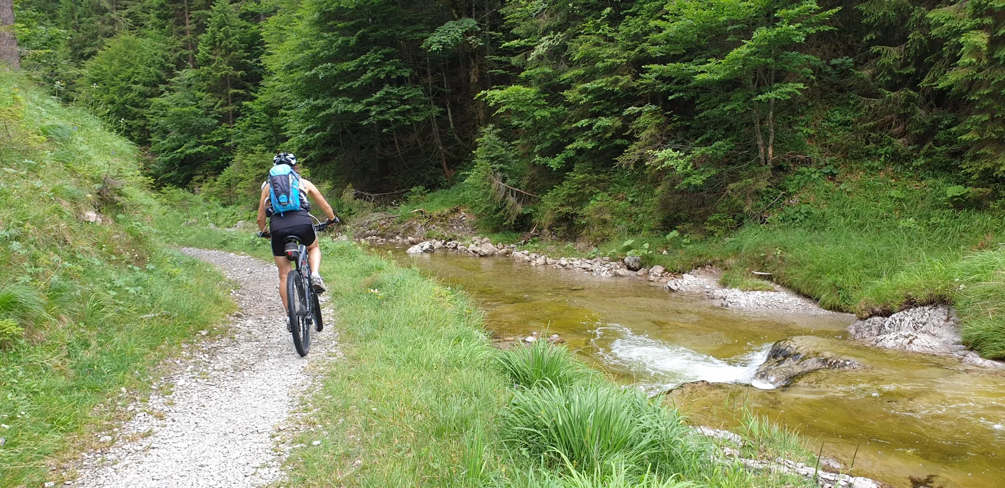Auf einem Schotterweg an einem Bergbach entlang fährt ein Mountainbiker vom Betrachter weg