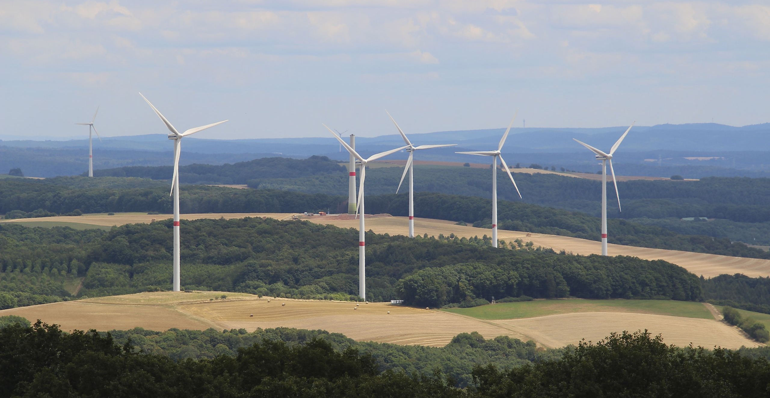 Blick aus der Luft über eine hügelige Wald-Feld-Landschaft, in der im Vordergrund 6 Windräder stehen