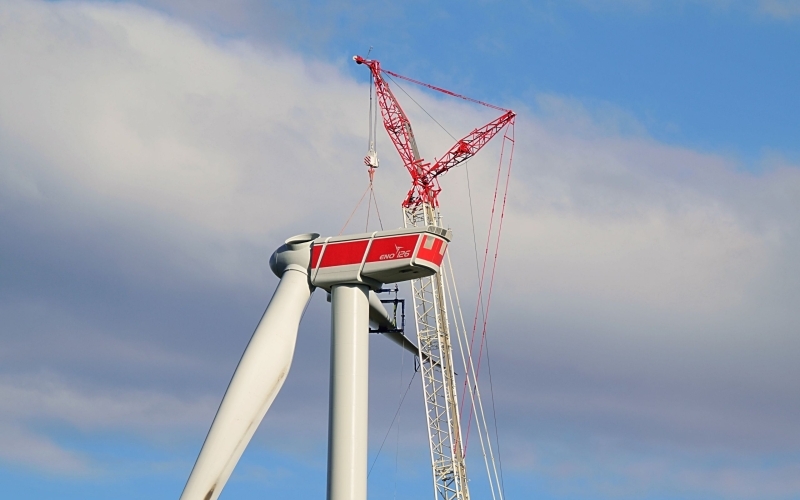 Blick auf die unfertige, rot-weiße Kanzel eines Windrads, an der ein Kran steht, der einen Rotor befestigt
