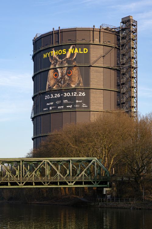 An einem hohen, zylindrischen Industriebau prangt ein Plakat mit Werbung für "Mythos Wald"