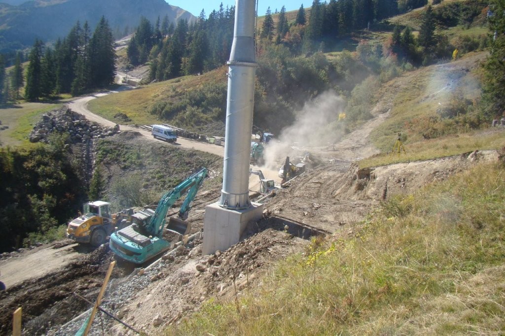 Blick auf eine Baustelle an einem Berghang, wo im Vordergrund ein massiver Mast gesetzt wurde, Bagger arbeiten und im Hintergrund ein Fahrzeug eine gut ausgebaute Trasse entlang gefahren kommt. 
