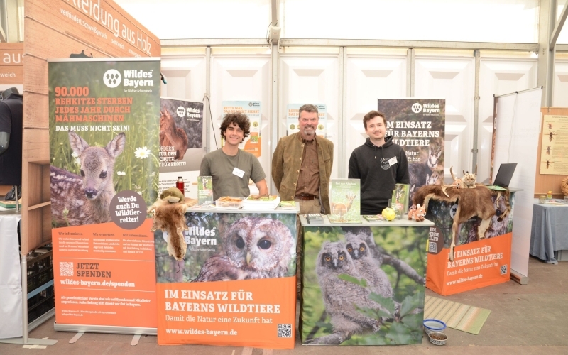 Hinter einem Messestand mit Naturmotiven und orangefarbenen Botschaften stehen drei Männer und lächeln freundlich in die Kamera