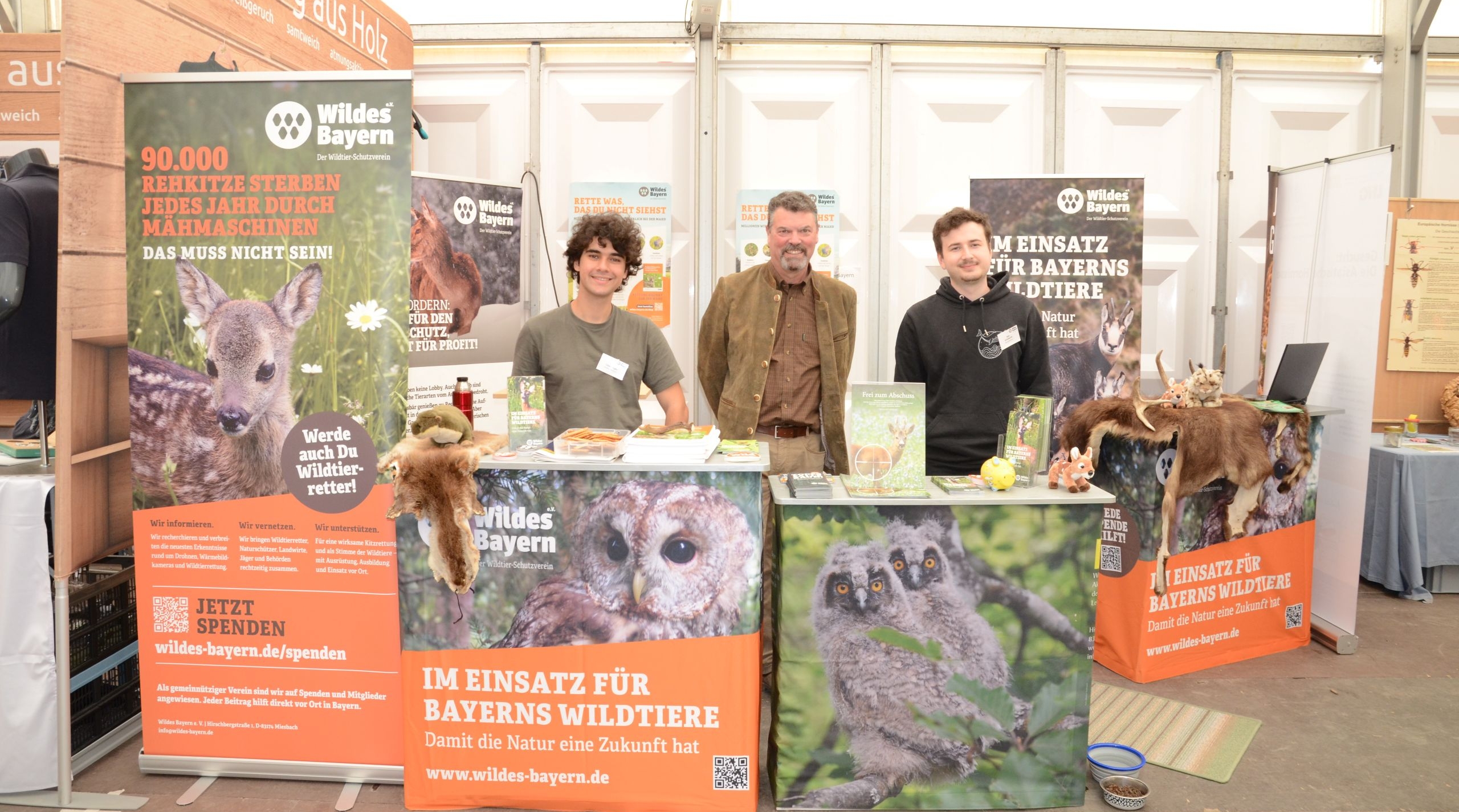 Hinter einem Messestand mit Naturmotiven und orangefarbenen Botschaften stehen drei Männer und lächeln freundlich in die Kamera